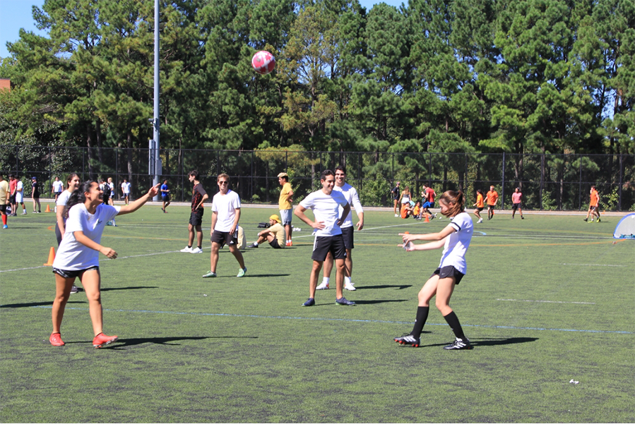 several students kick a soccer ball back and forth on a soccer field, with several other people in the background